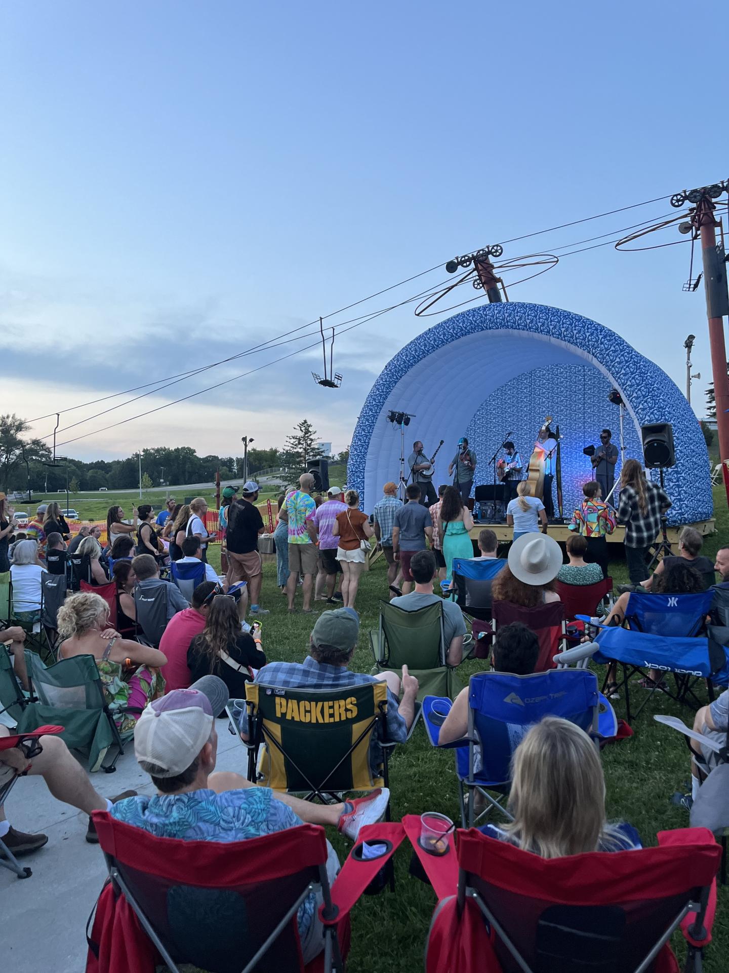 Outdoor concert with crowd seated on grass, stage in background at dusk.