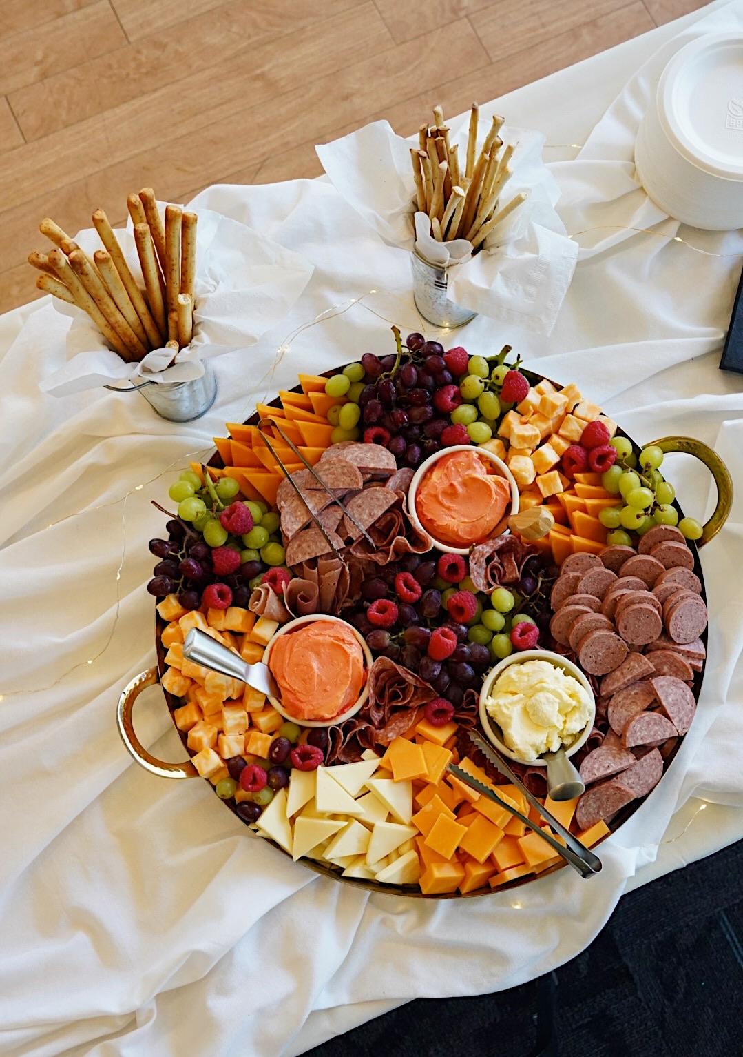 Cheese and meat platter with grapes, sauces, and pretzel sticks on a white cloth.