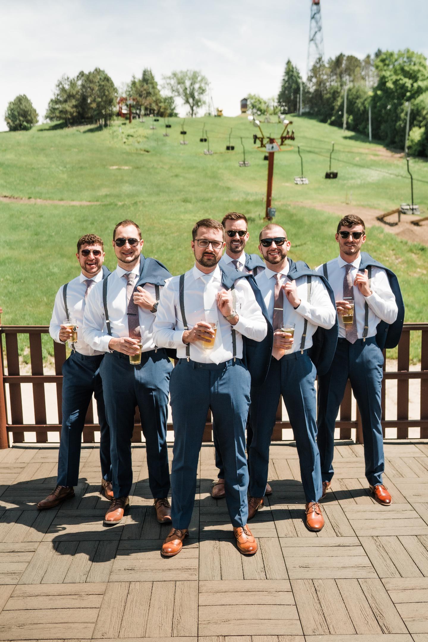 Groom and groomsmen in blue suits, holding drinks on a sunny day with a green hill behind.