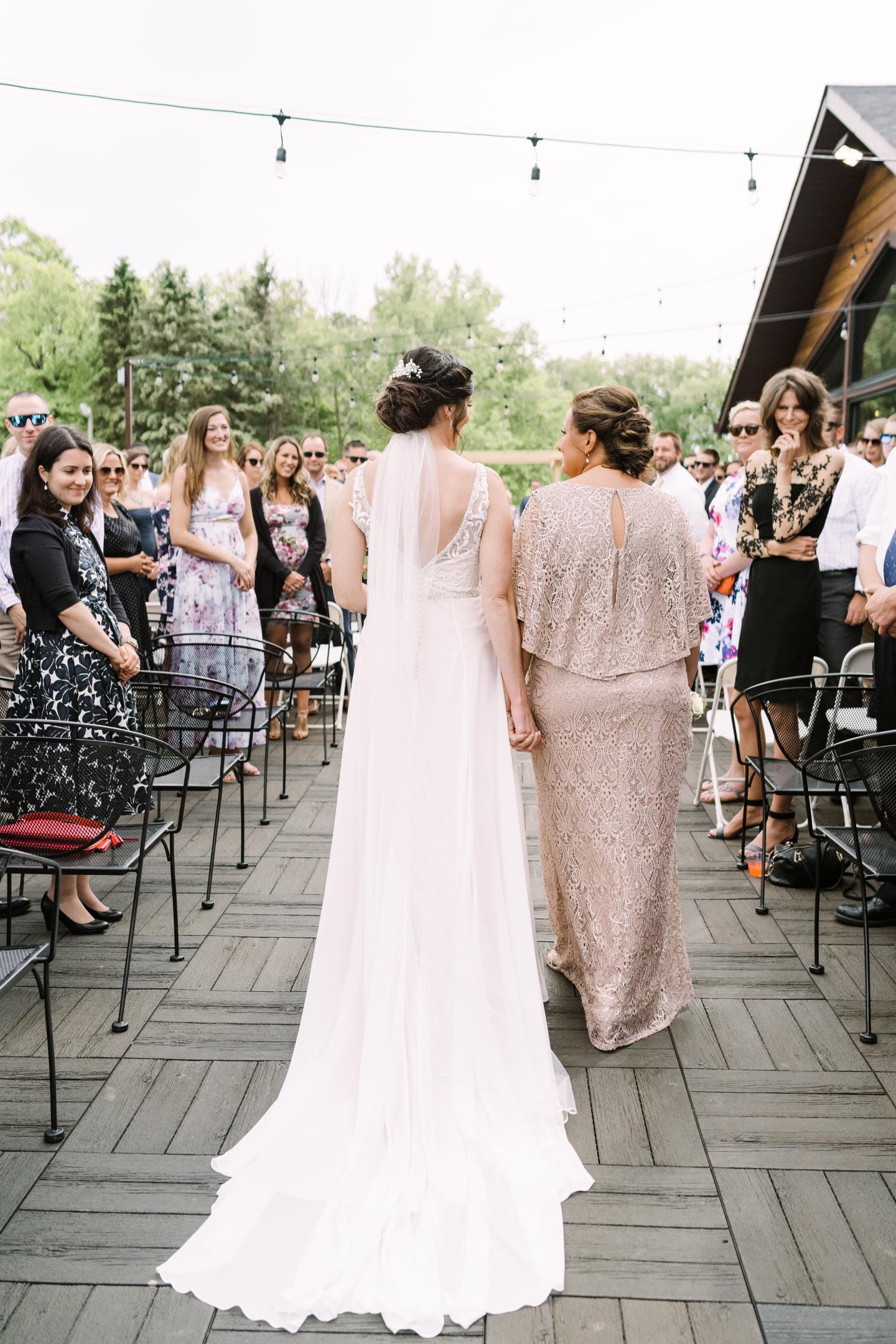 Bride and woman in beige walking down an outdoor aisle, guests seated on either side.