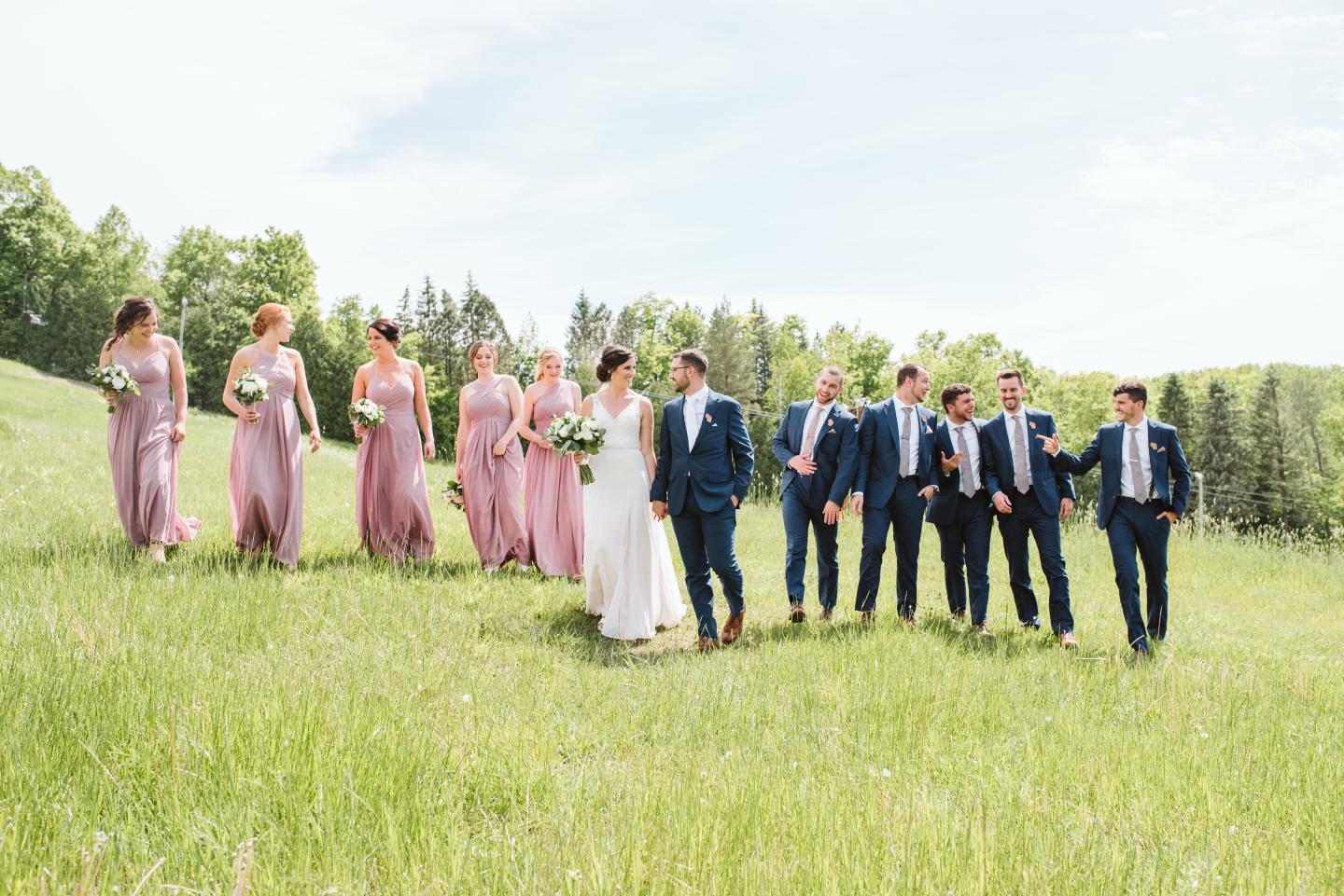 Bridal party walking on a grassy field in suits and lilac dresses.