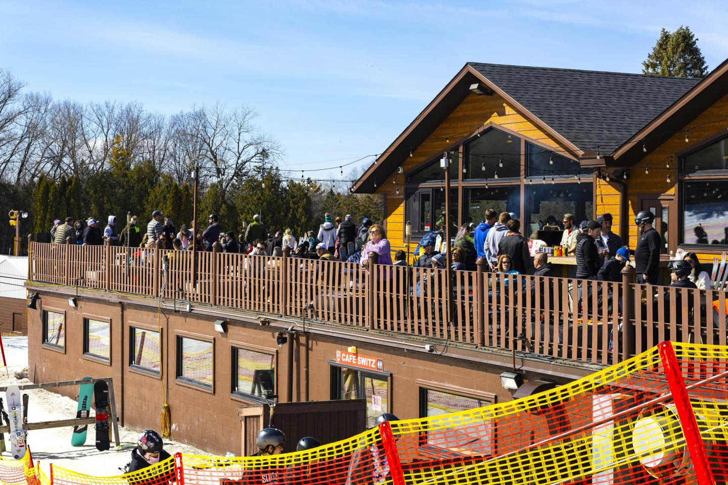 Ski lodge with a wooden deck crowded with people. Snow and trees in the background.