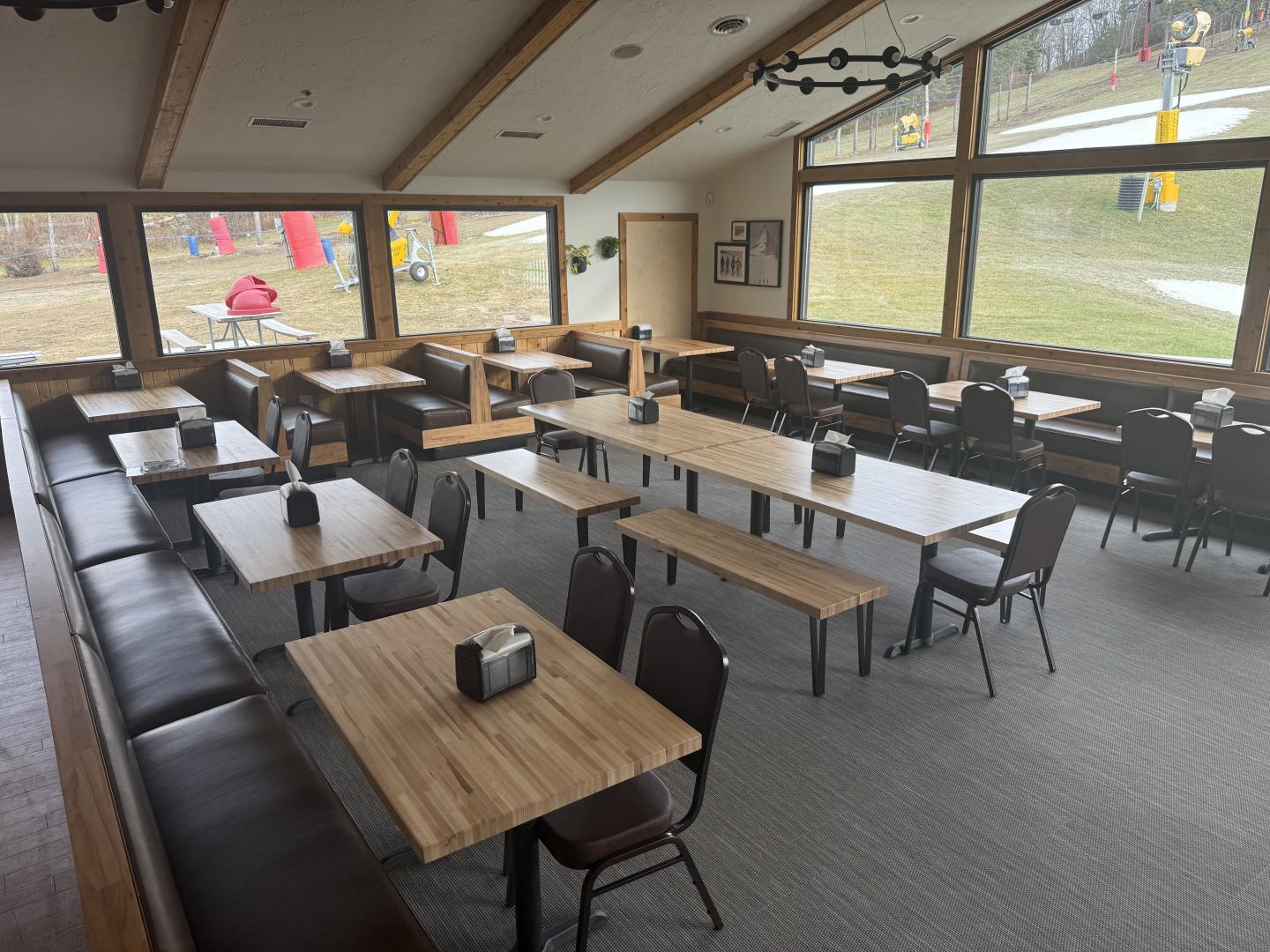 Empty dining area with wooden tables and chairs, large windows overlook a grassy outdoor area.