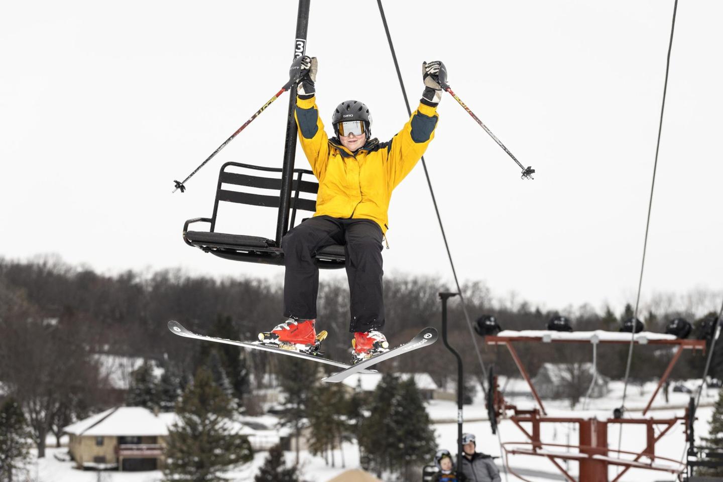 Skier on lift with arms raised, snowy backdrop.
