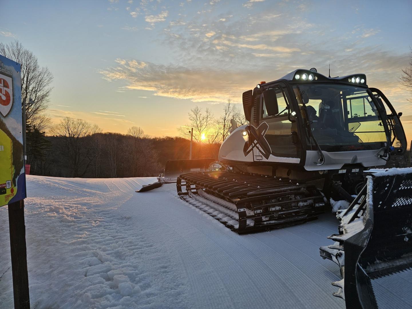 Snow grooming machine on a snowy slope at sunset.