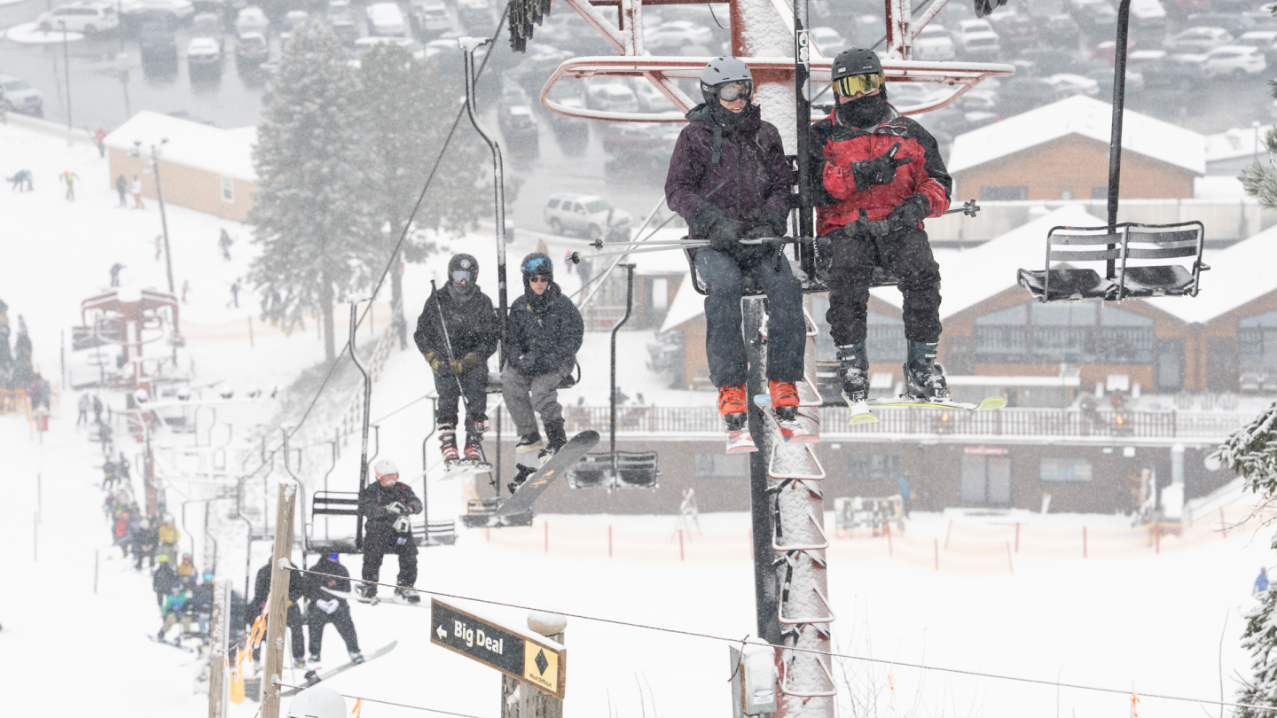 Skiers on a snowy mountain chairlift, with buildings in the background.