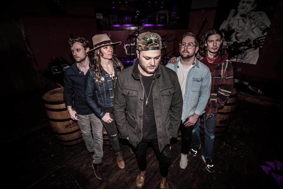 Five people standing in a dimly lit room, with a musician mural in the background.