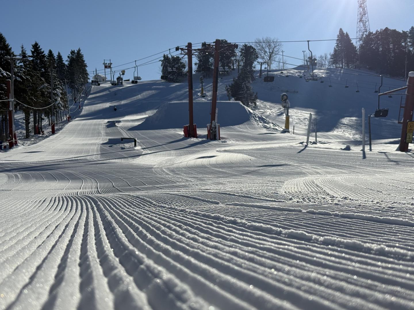 Groomed ski slope under clear blue sky, with trees and ski lift poles.