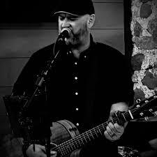 Musician sings and plays acoustic guitar, wearing a cap. Black and white photo.