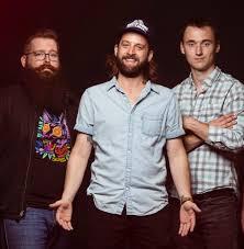 Three men standing, one with a beard and glasses, another with a cap, smiling.