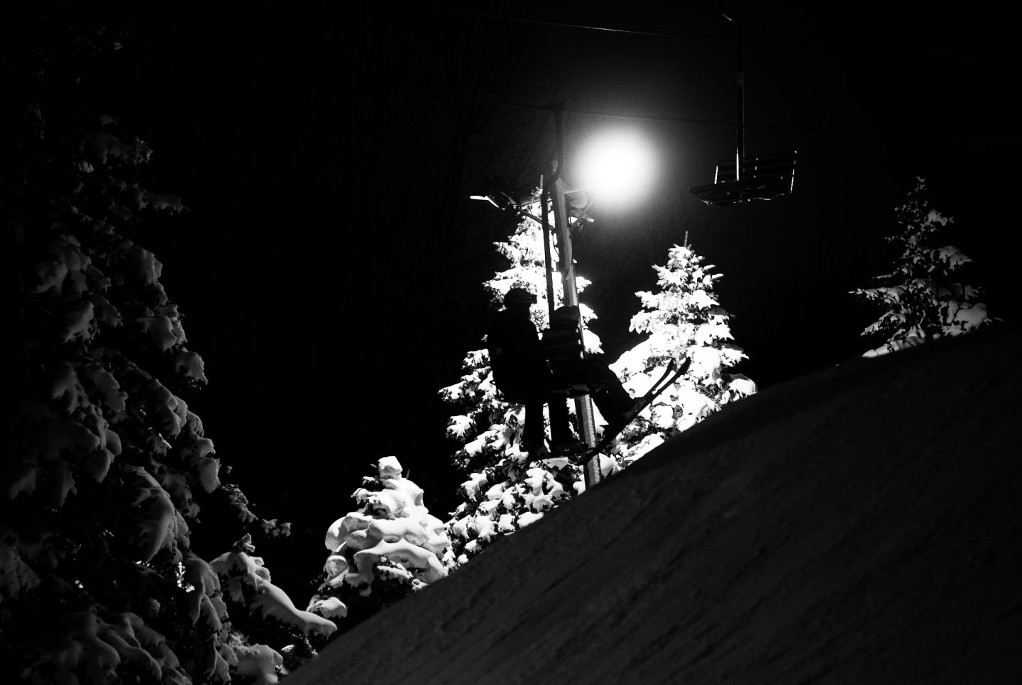 Silhouetted skier on chairlift at night, snowy trees lit in background.
