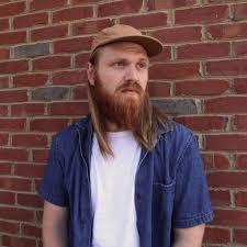 Man with long hair, beard, cap, denim shirt, leaning on brick wall.