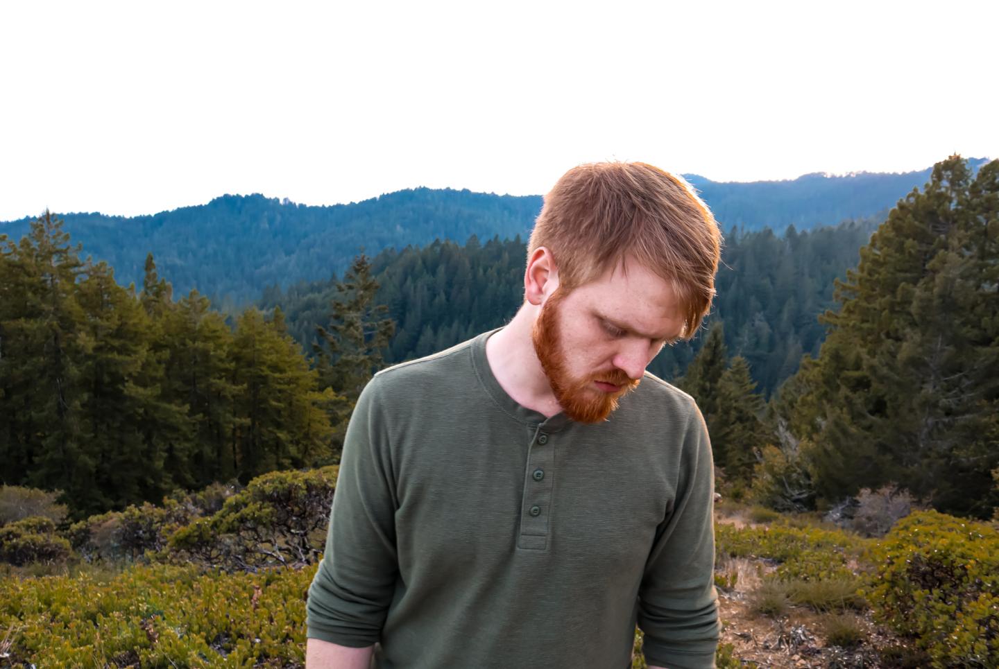 A person with a beard looks down in a forest with hills in the background.