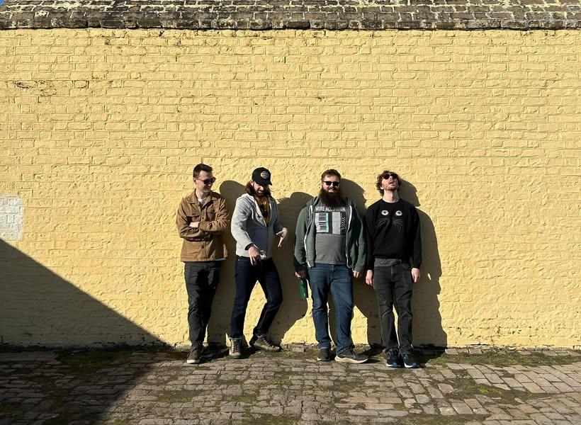 Four people stand against a sunlit yellow brick wall.
