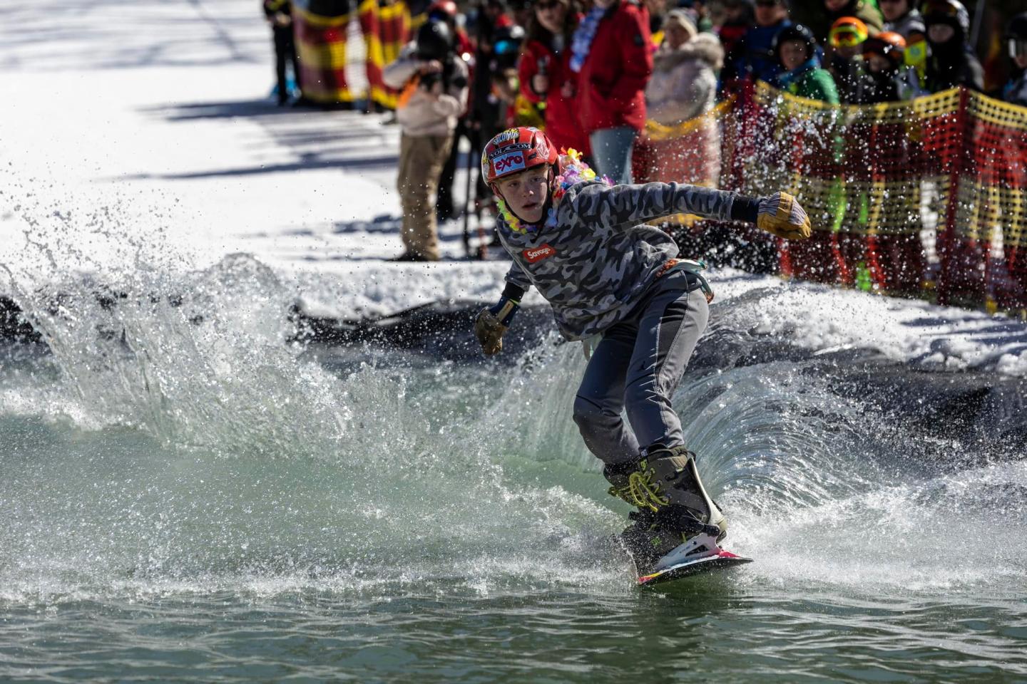 Snowboarder glides across a pool, splashing water, with a crowd watching.