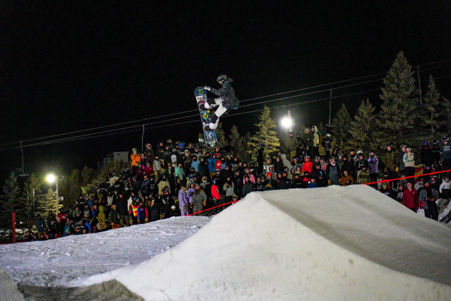 Snowboarder in mid-air at night event, crowd watching.