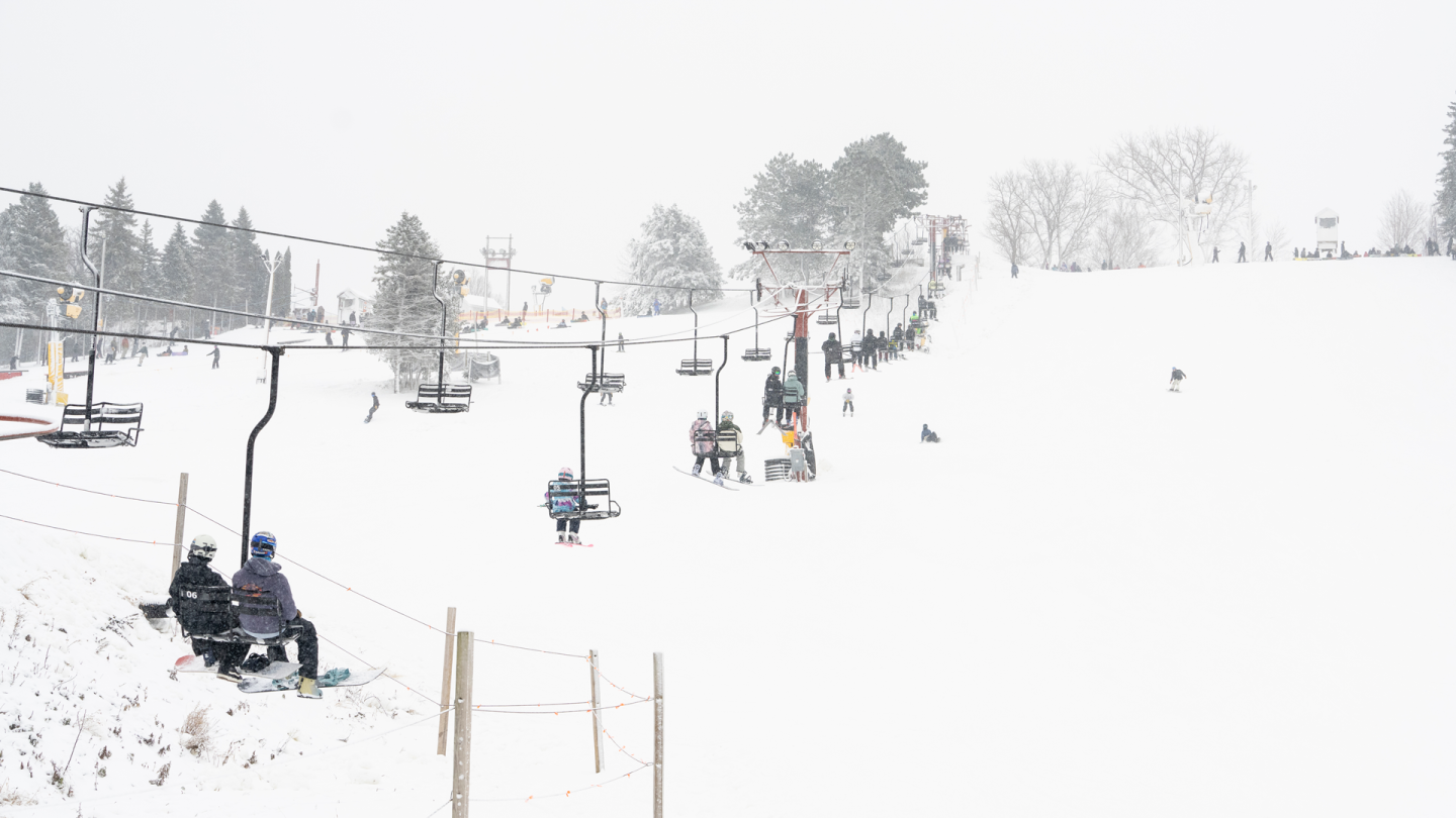 Skiers on a snowy slope with a chairlift and trees in the background.