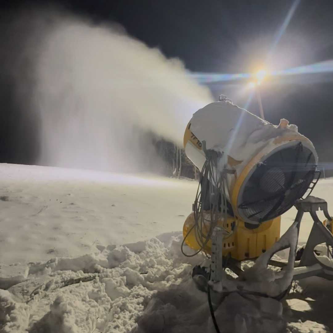 Snow machine blowing snow under bright lights at night.