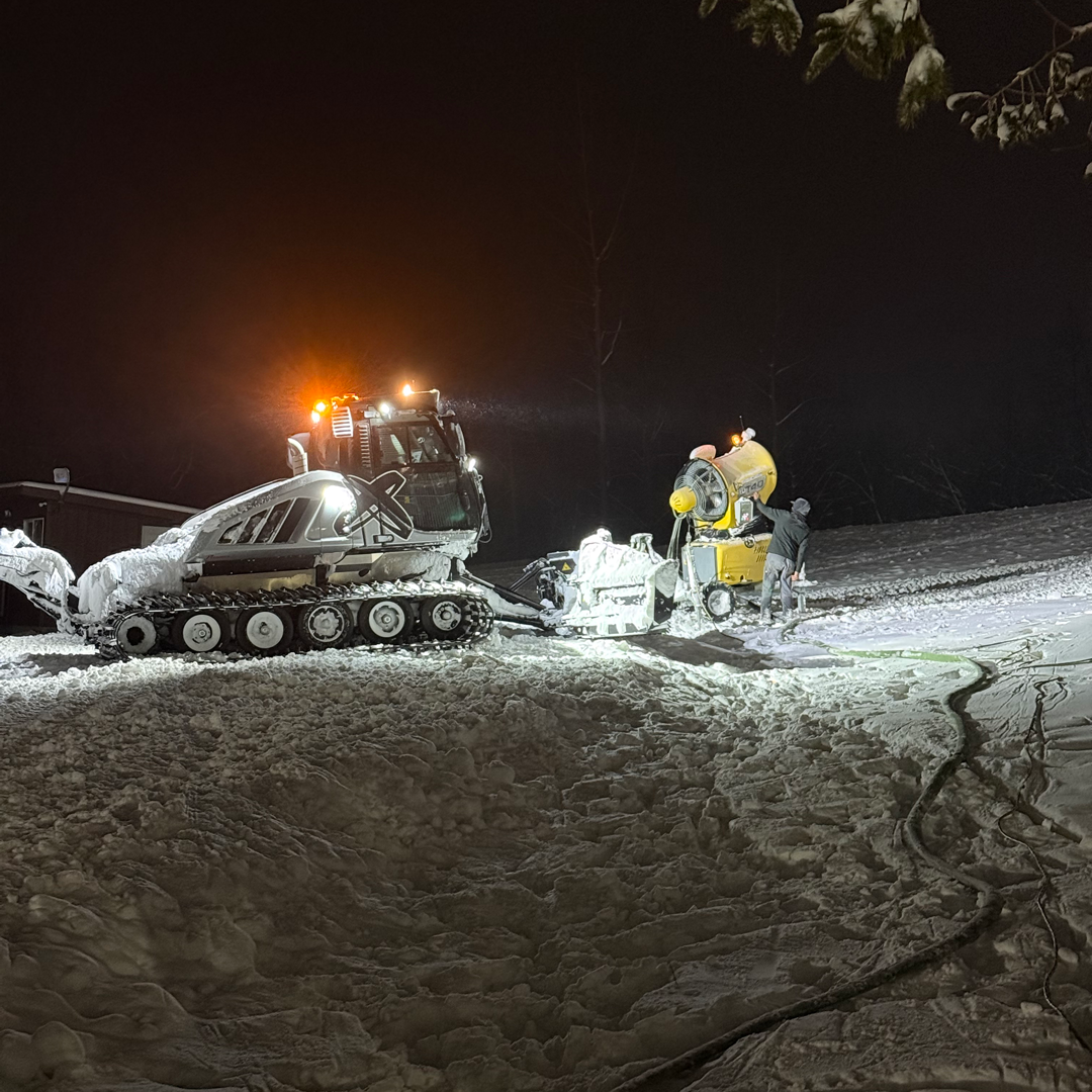 Snowgroomer and workers making snow on a slope at night, bright lights illuminating.