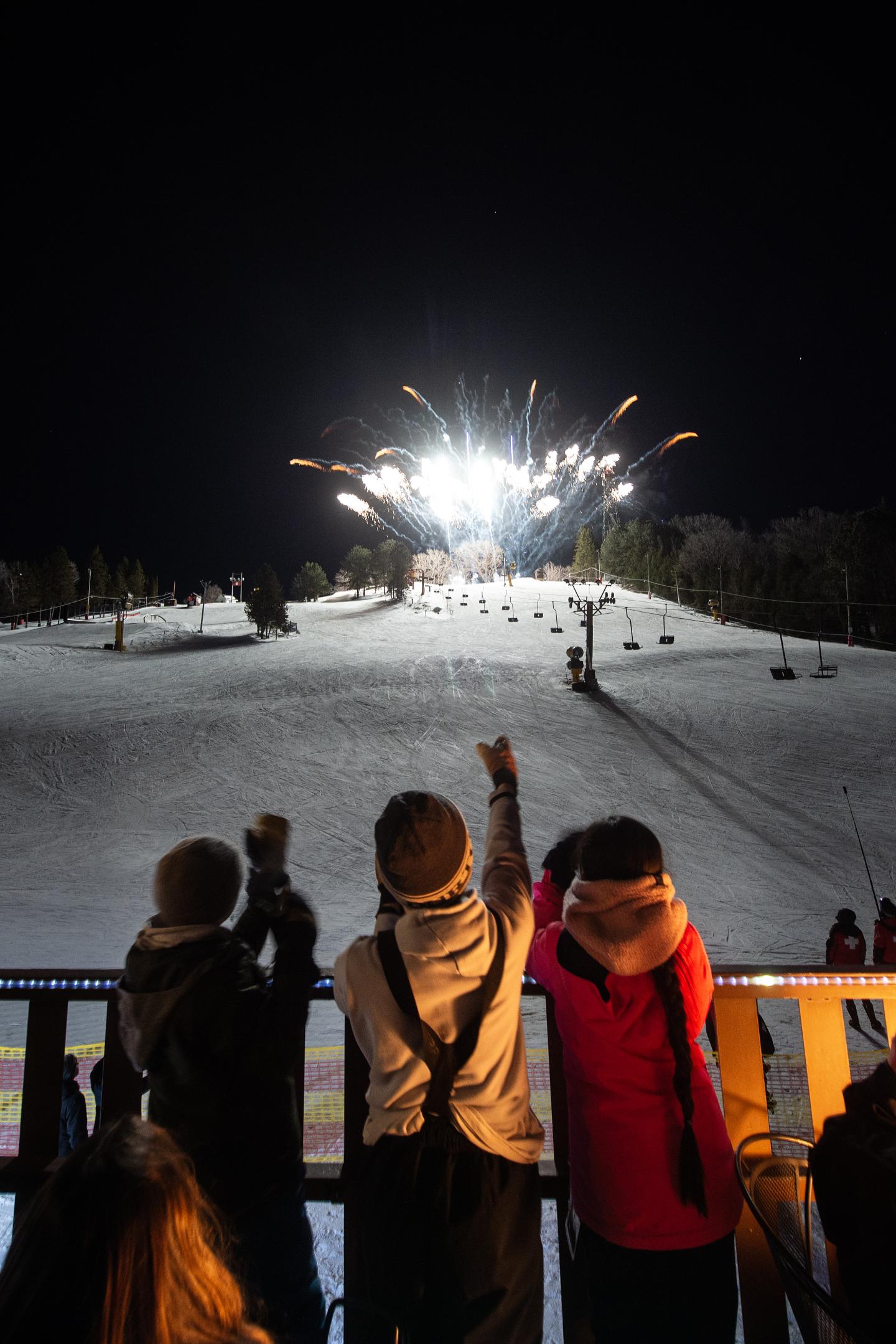 Kids watching fireworks over snowy hill at night.