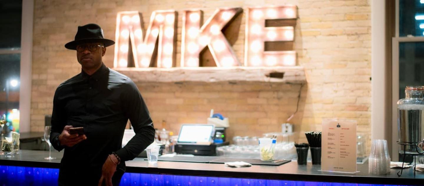Man in dark clothing leaning on a bar under illuminated MKE sign.