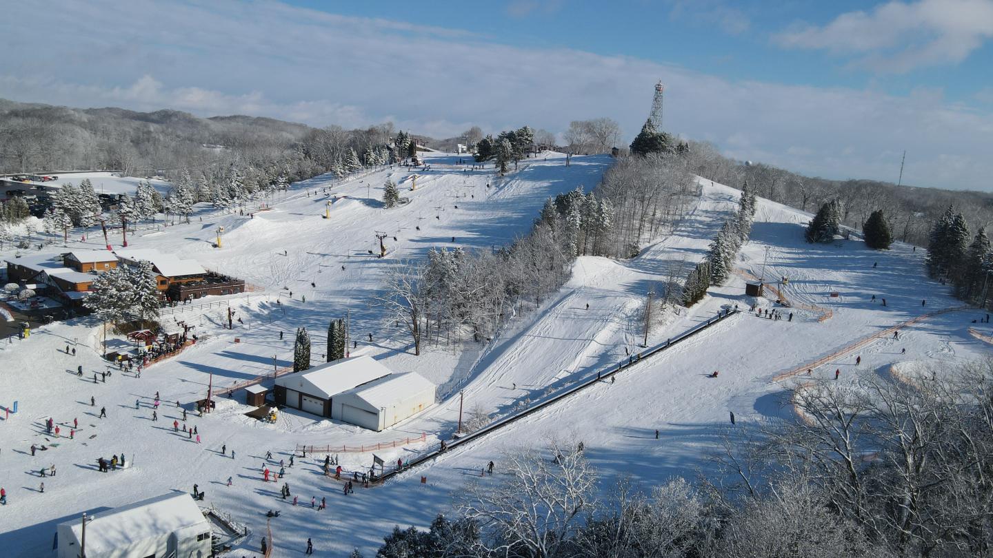 Snowy ski resort with skiers, trees, and distant statue under a blue sky.