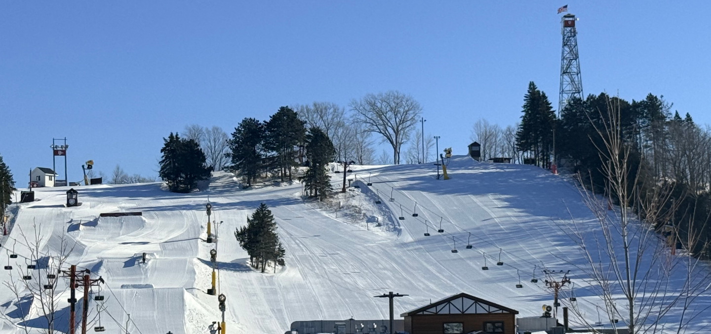 Snow-covered ski slope with trees and a tower under a clear blue sky.