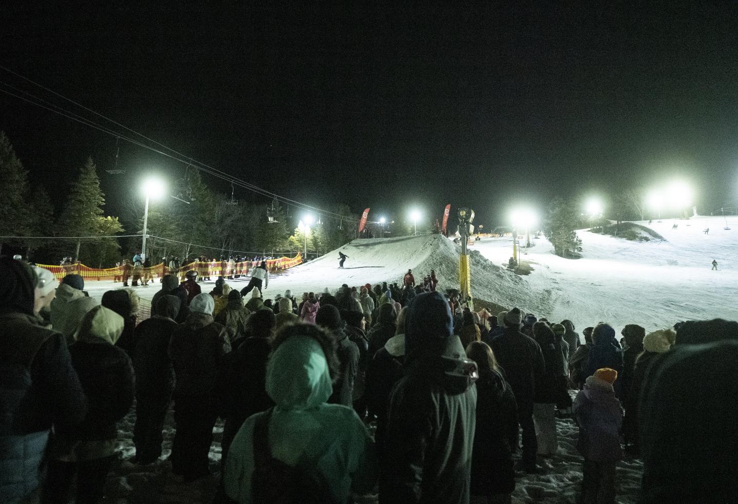 Crowd watching snowy hillside at night under bright lights.
