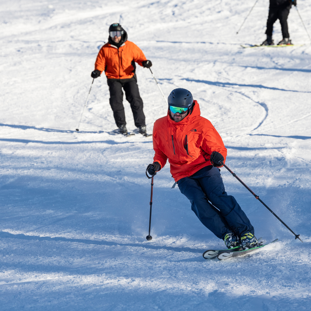 Two skiers in orange jackets on a snowy slope, skiing downhill.