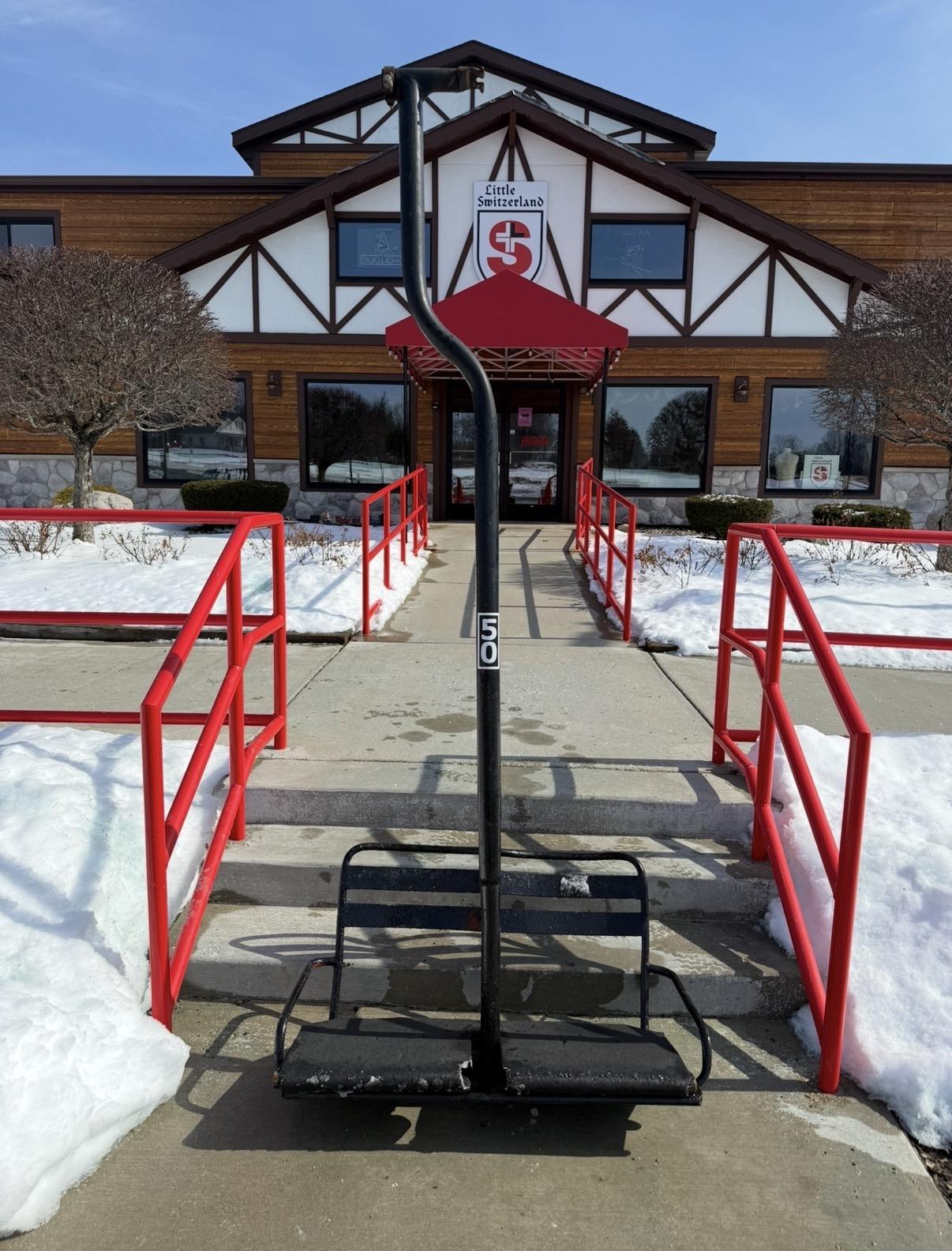 Ski lift chair on snowy path leading to a lodge with red railings.