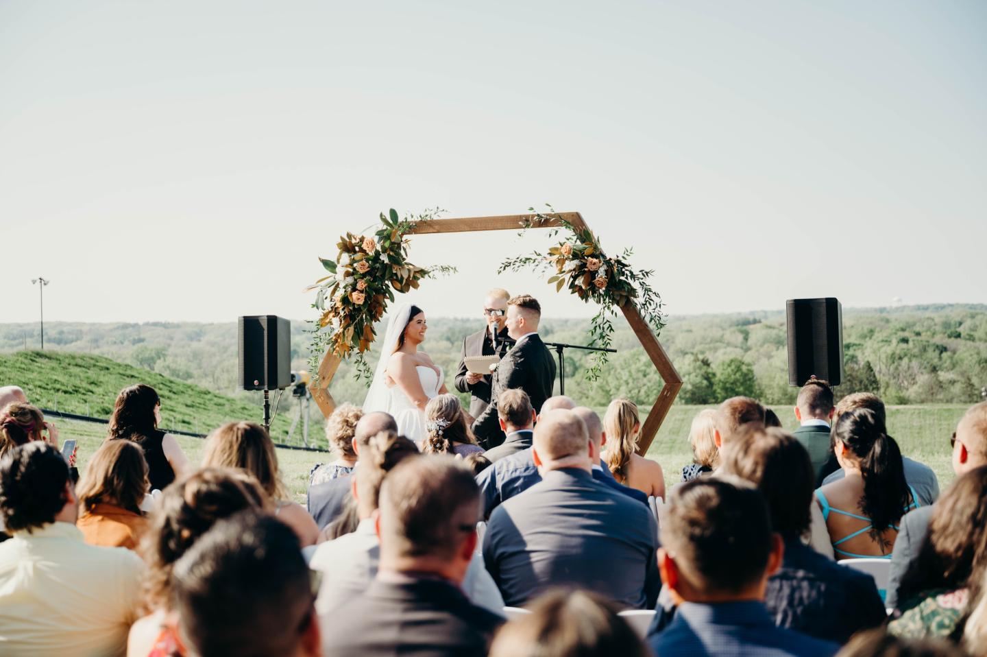 Outdoor wedding ceremony with a couple under a floral arch, guests seated.
