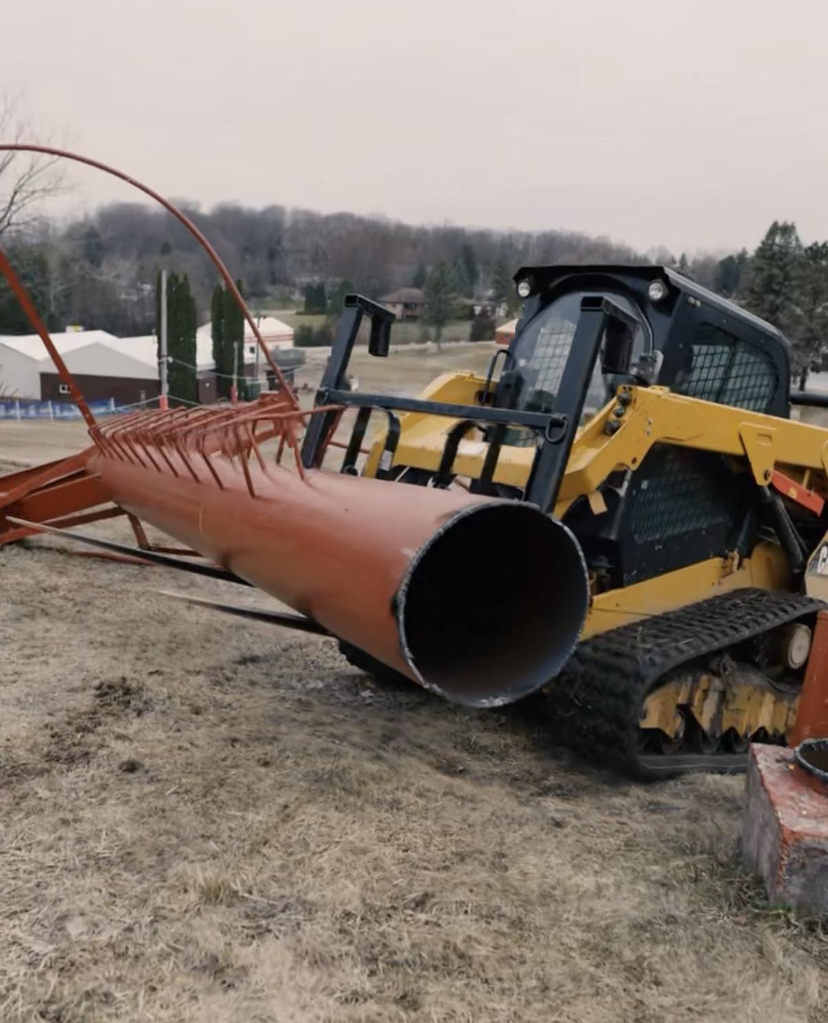 Bulldozer moving a large red pipe on a grassy field.