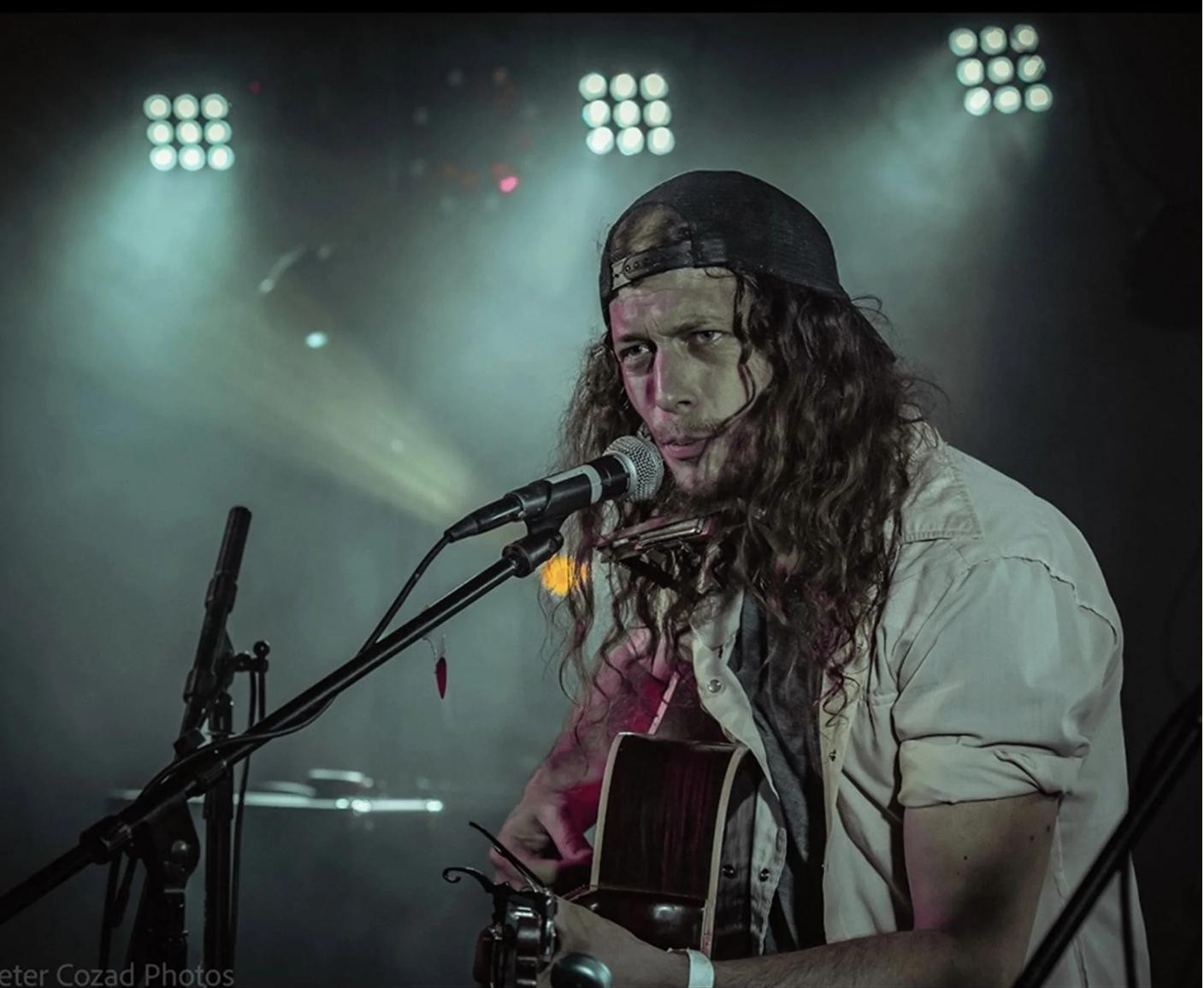 Musician with long hair playing guitar on stage under spotlights.