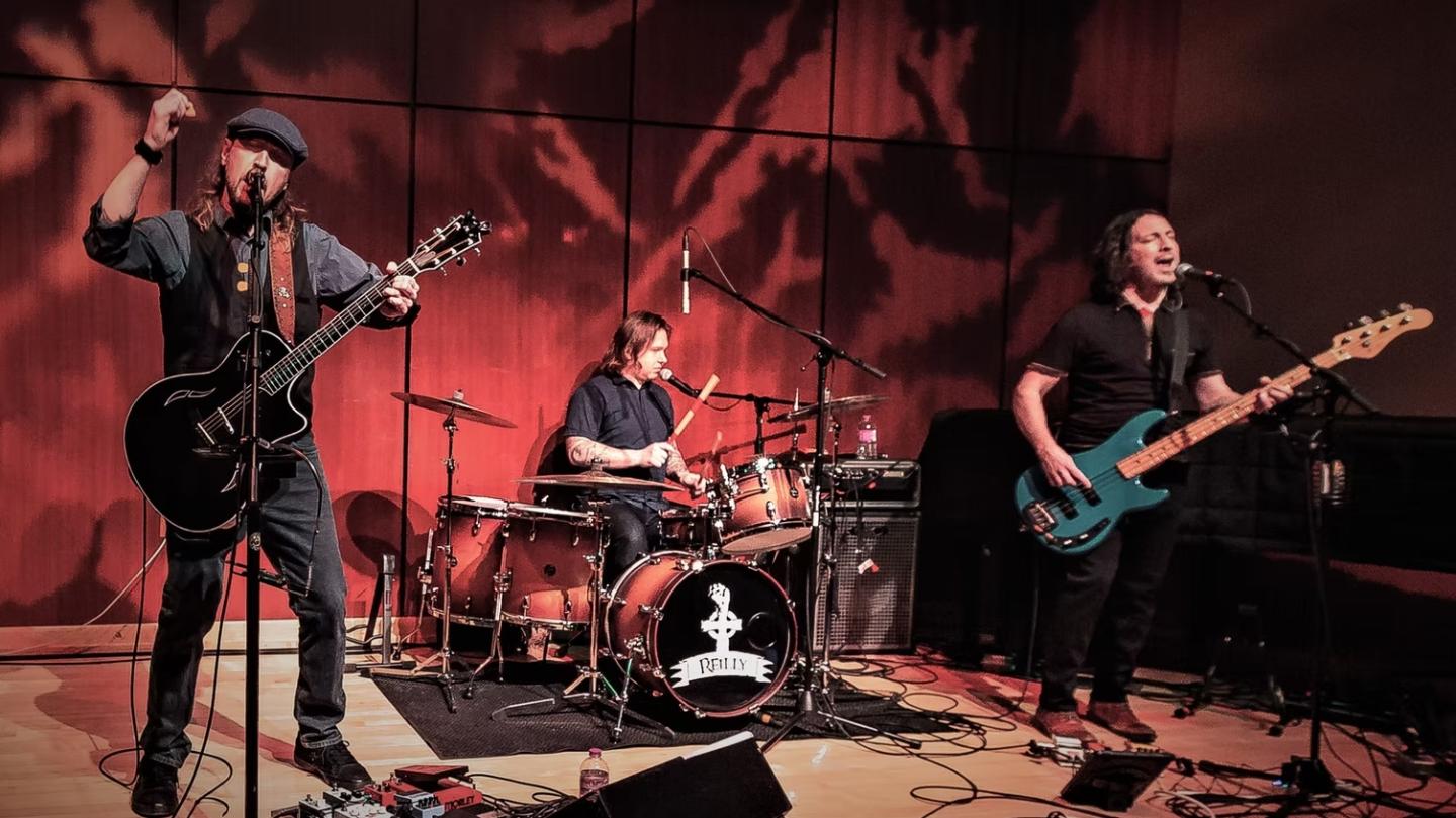 Three musicians performing on stage with guitar, drums, and bass.