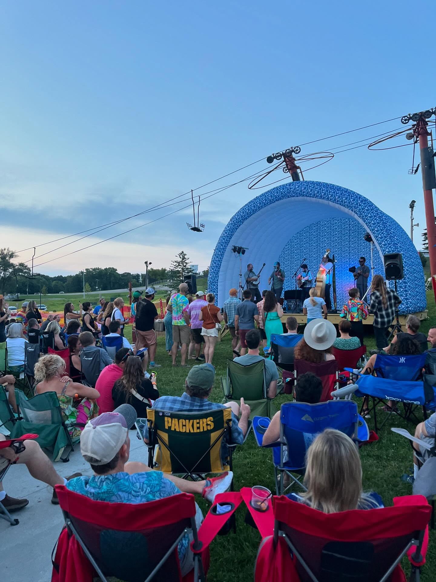 Outdoor concert audience sitting in chairs, facing a band on a domed stage at dusk.