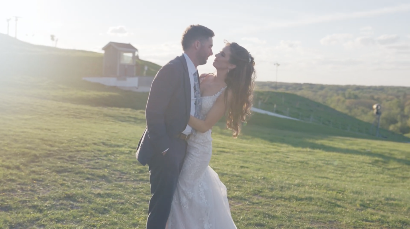 Couple embracing on a sunny hillside, dressed in wedding attire.