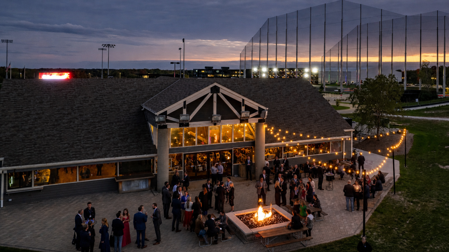 Outdoor evening event with people gathered around a fire pit, festive lights overhead.