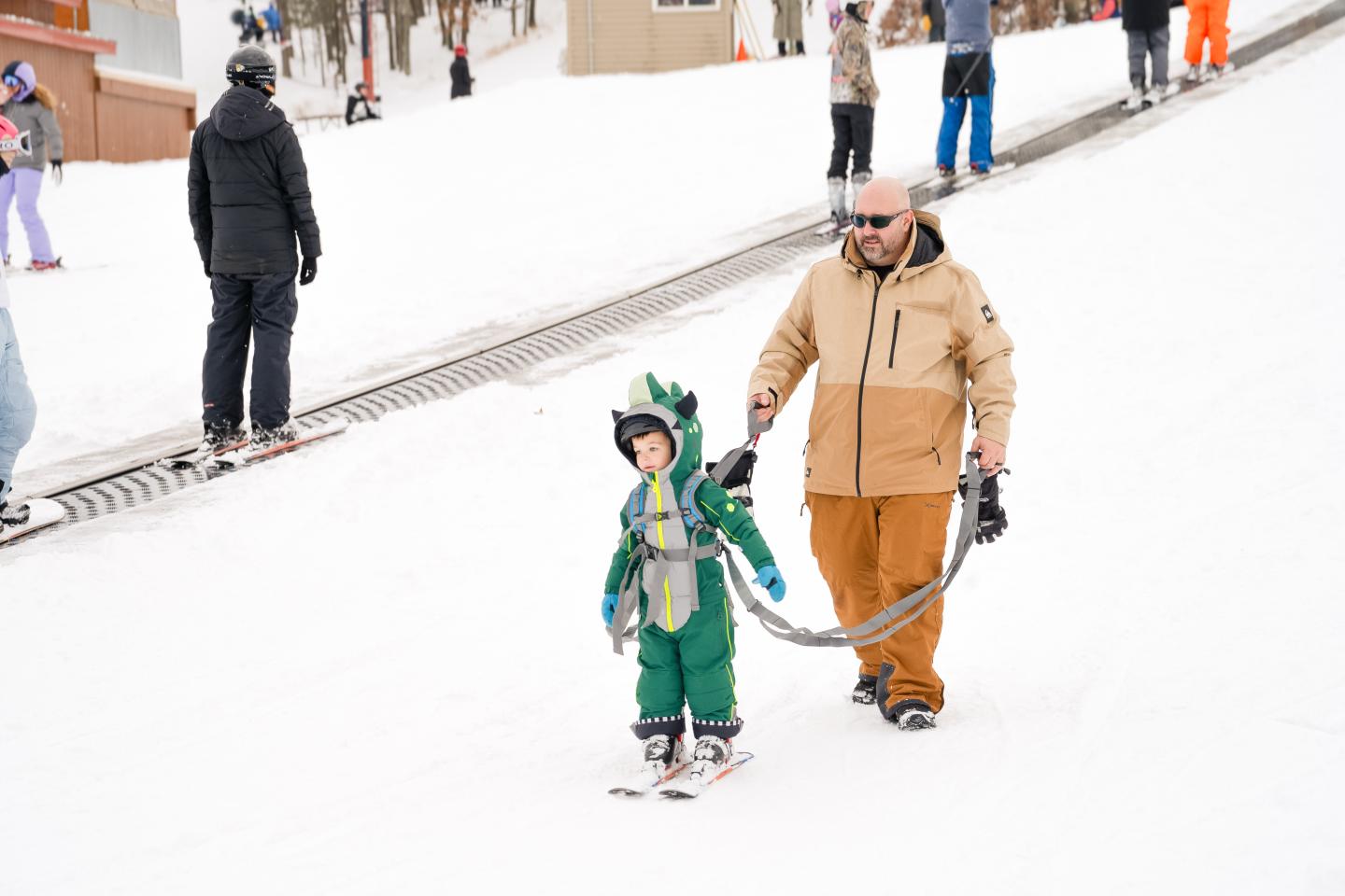 Child in green snowsuit skiing, guided by adult wearing a beige jacket on a snowy slope.