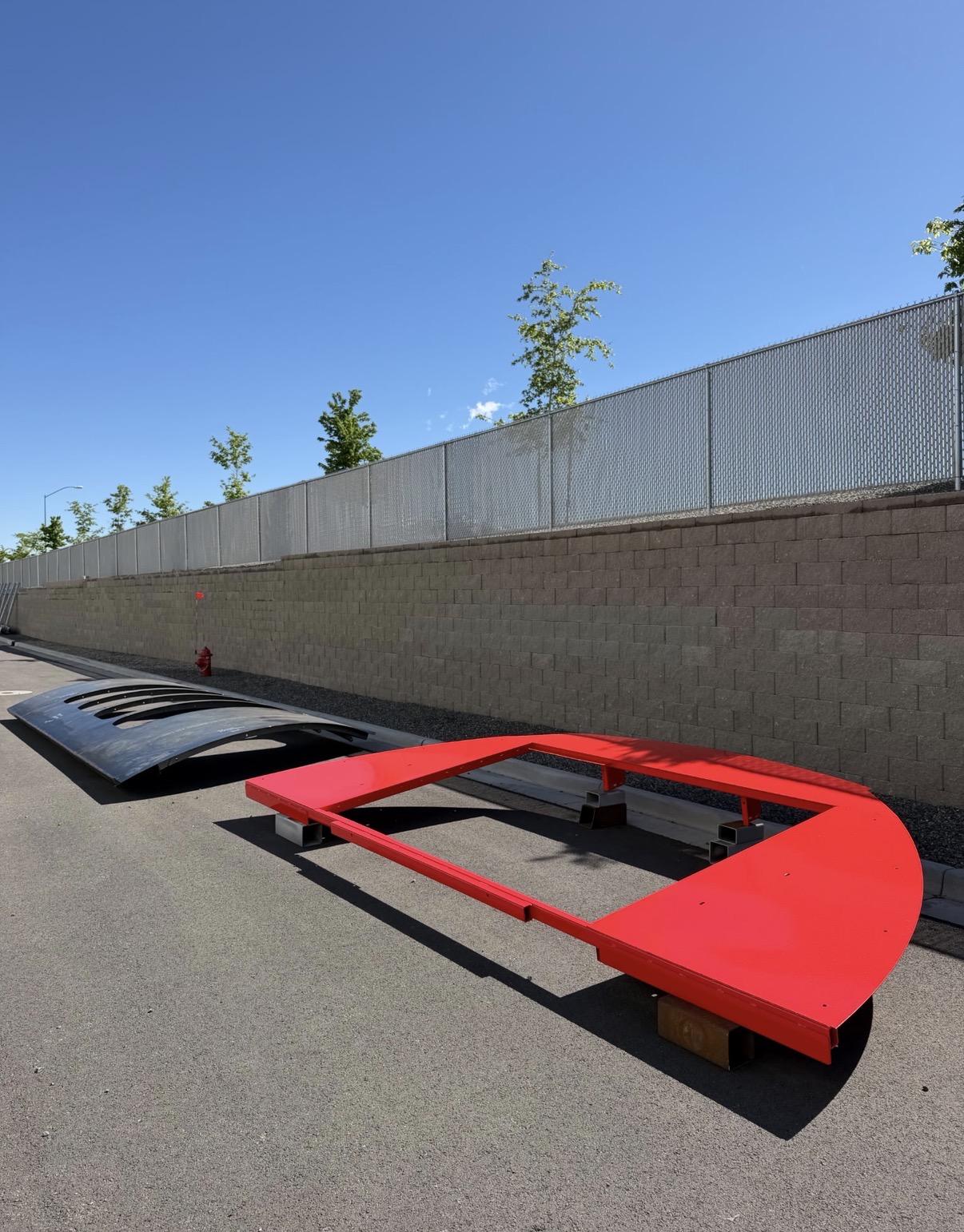 Red and black metal structures on a sunny road beside a brick wall.