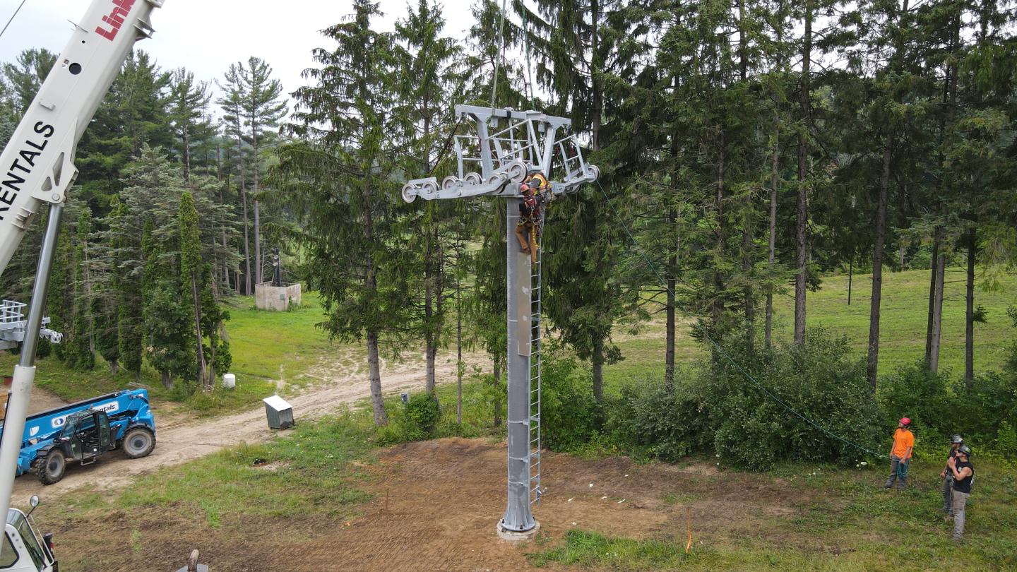 Workers installing chairlift in a forest clearing, with crane and safety gear.
