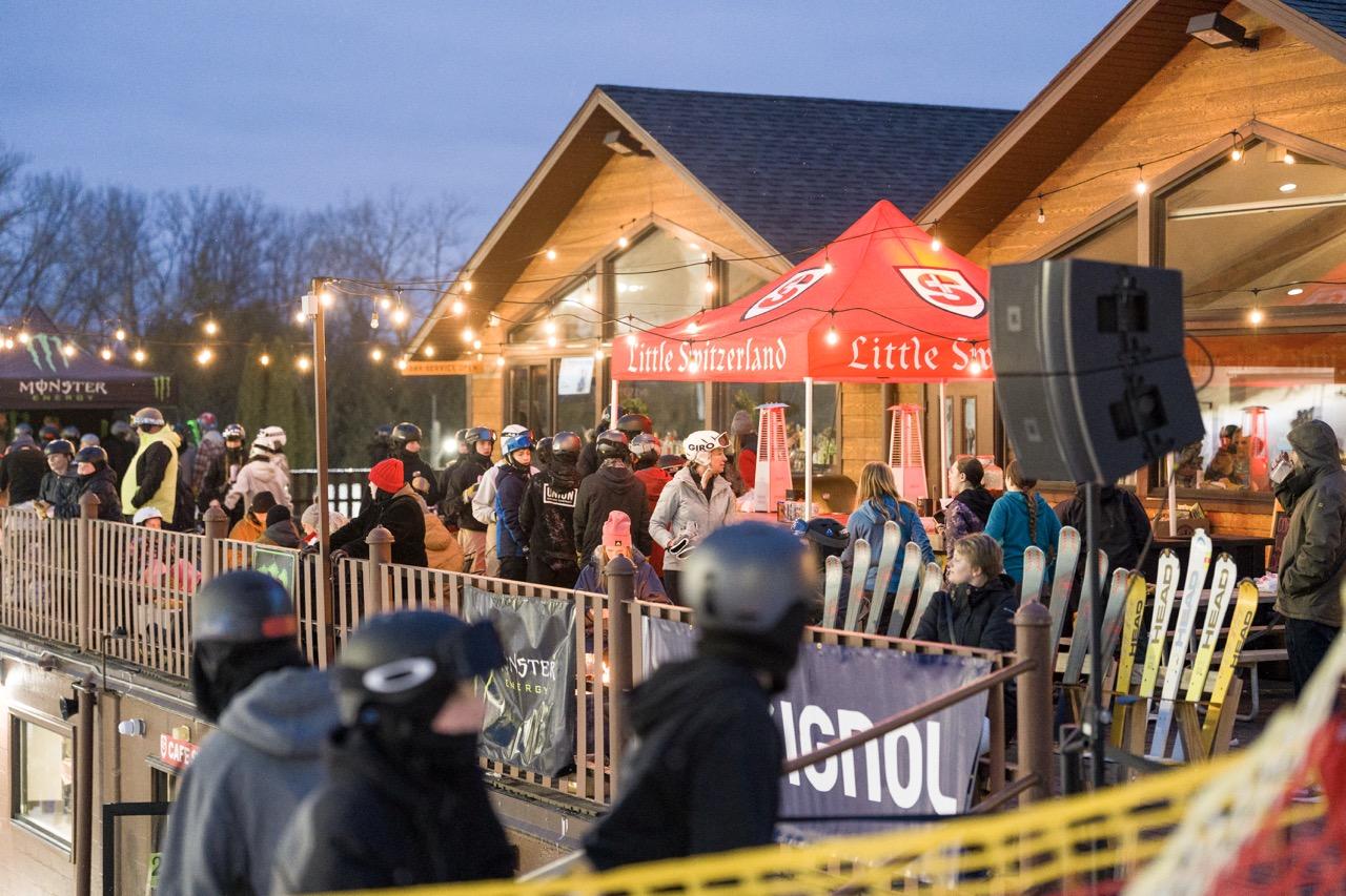 Crowd gathers at a ski rental booth outside a warmly lit lodge at dusk.