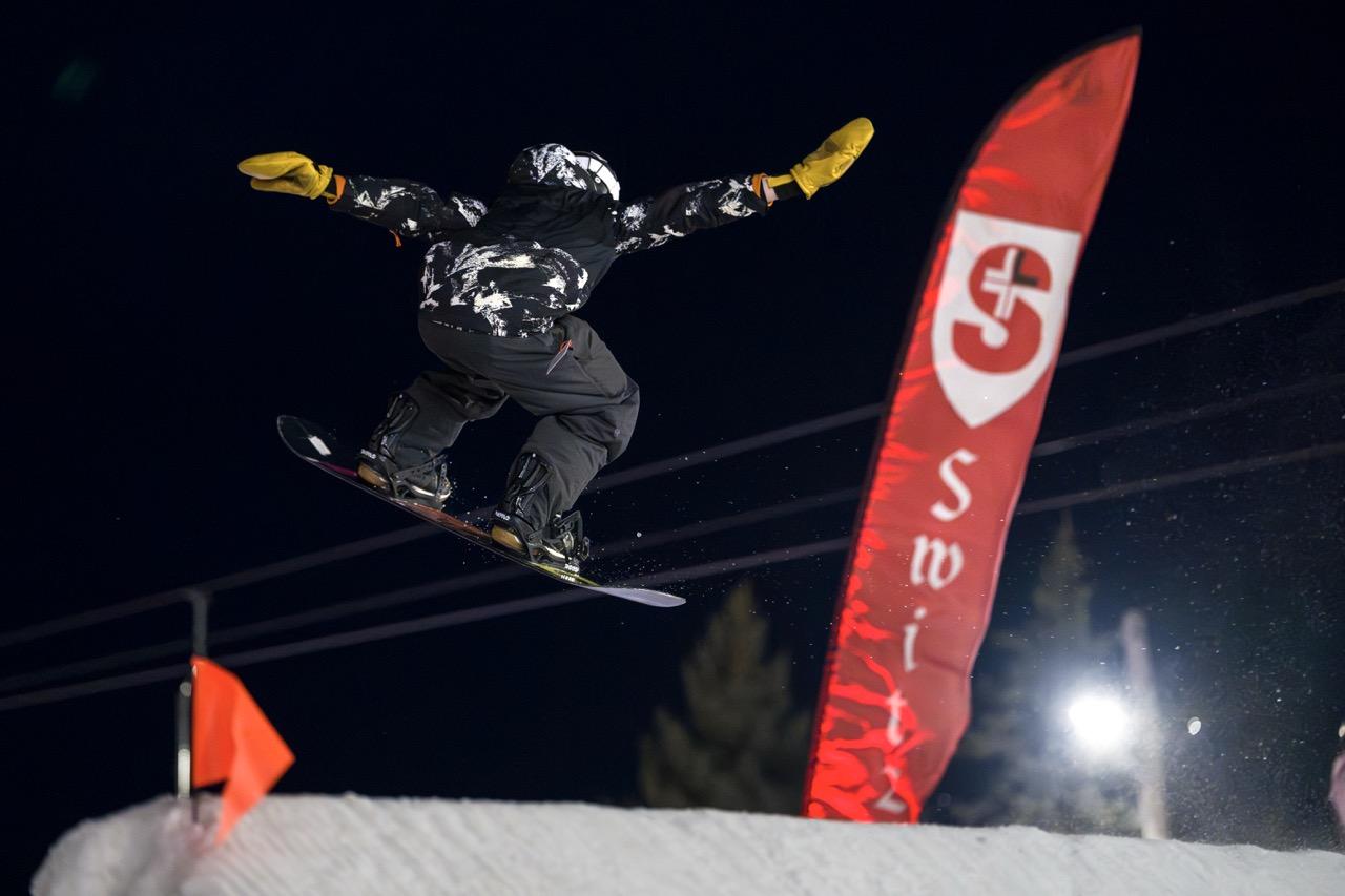 Snowboarder airborne over a rail at night beside a red sponsor flag
