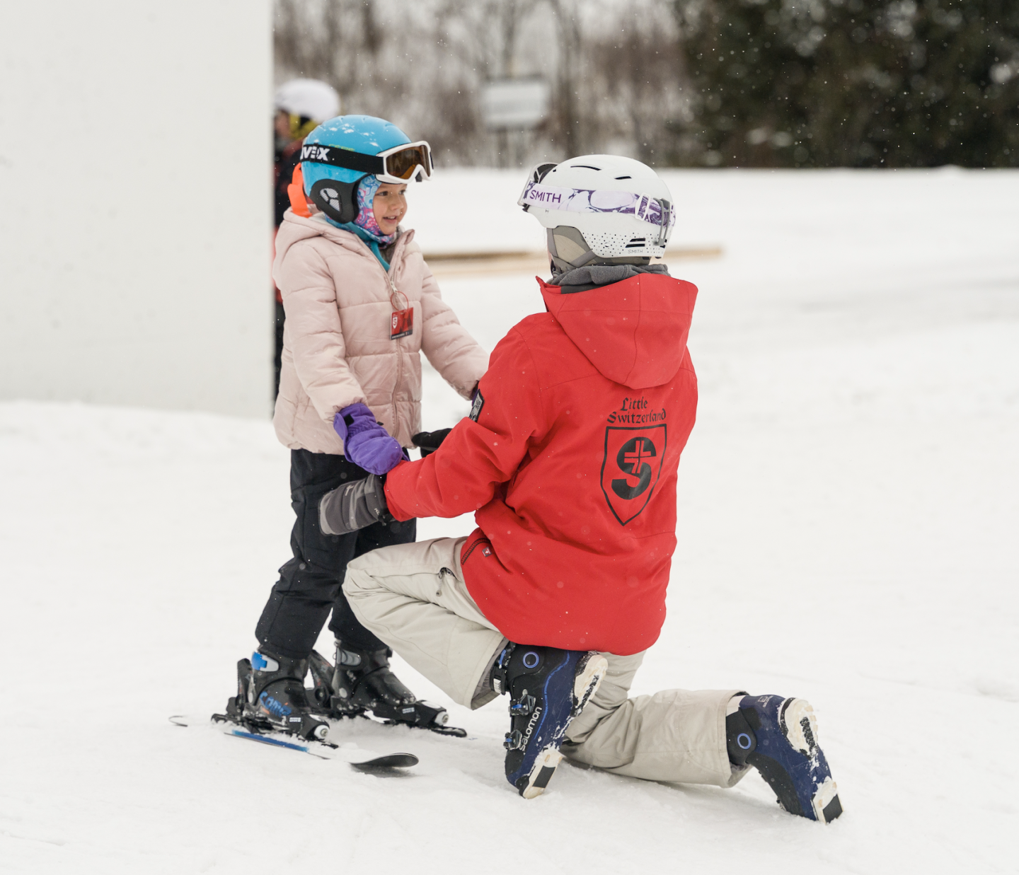 Adult kneeling in snow helps a child wearing skis and a helmet.