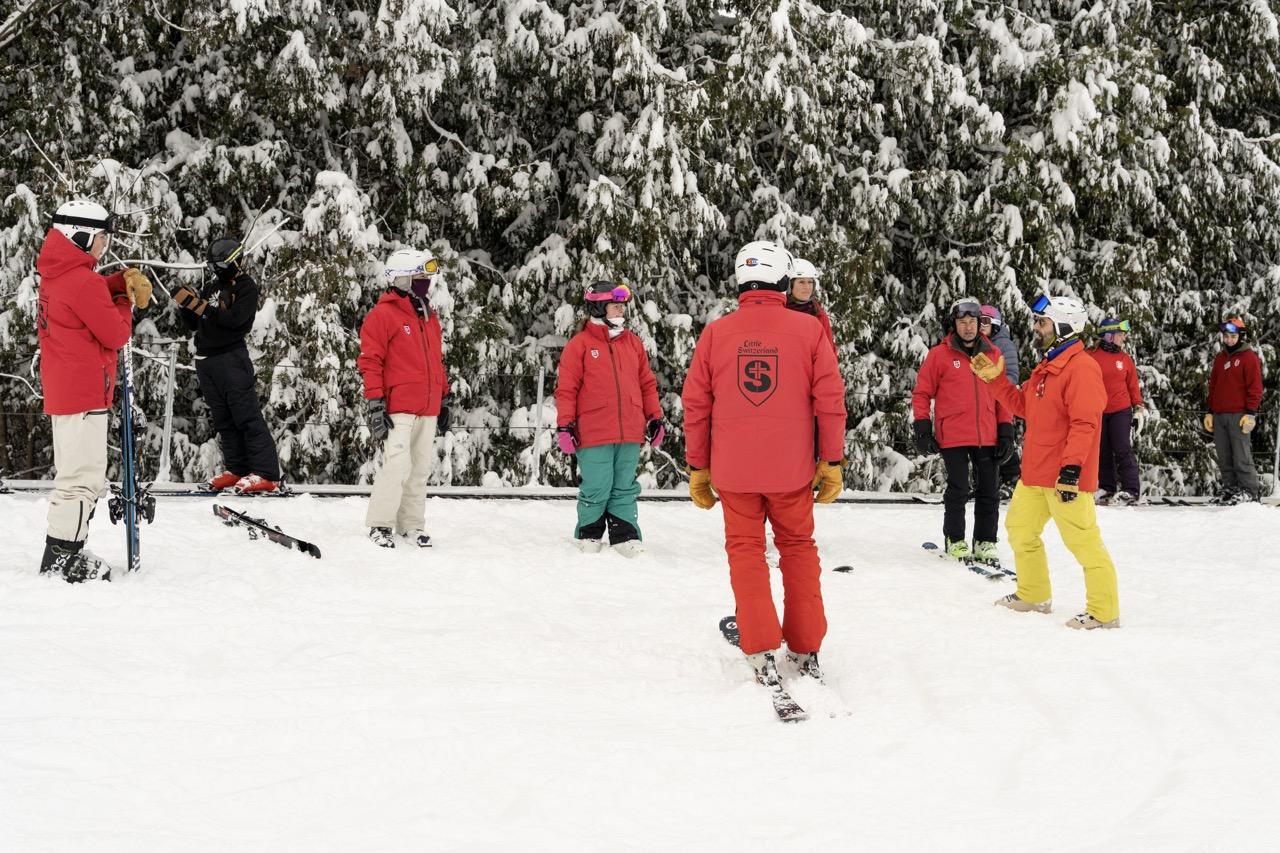 Skiers in colorful jackets gather on a snowy slope before a forest.