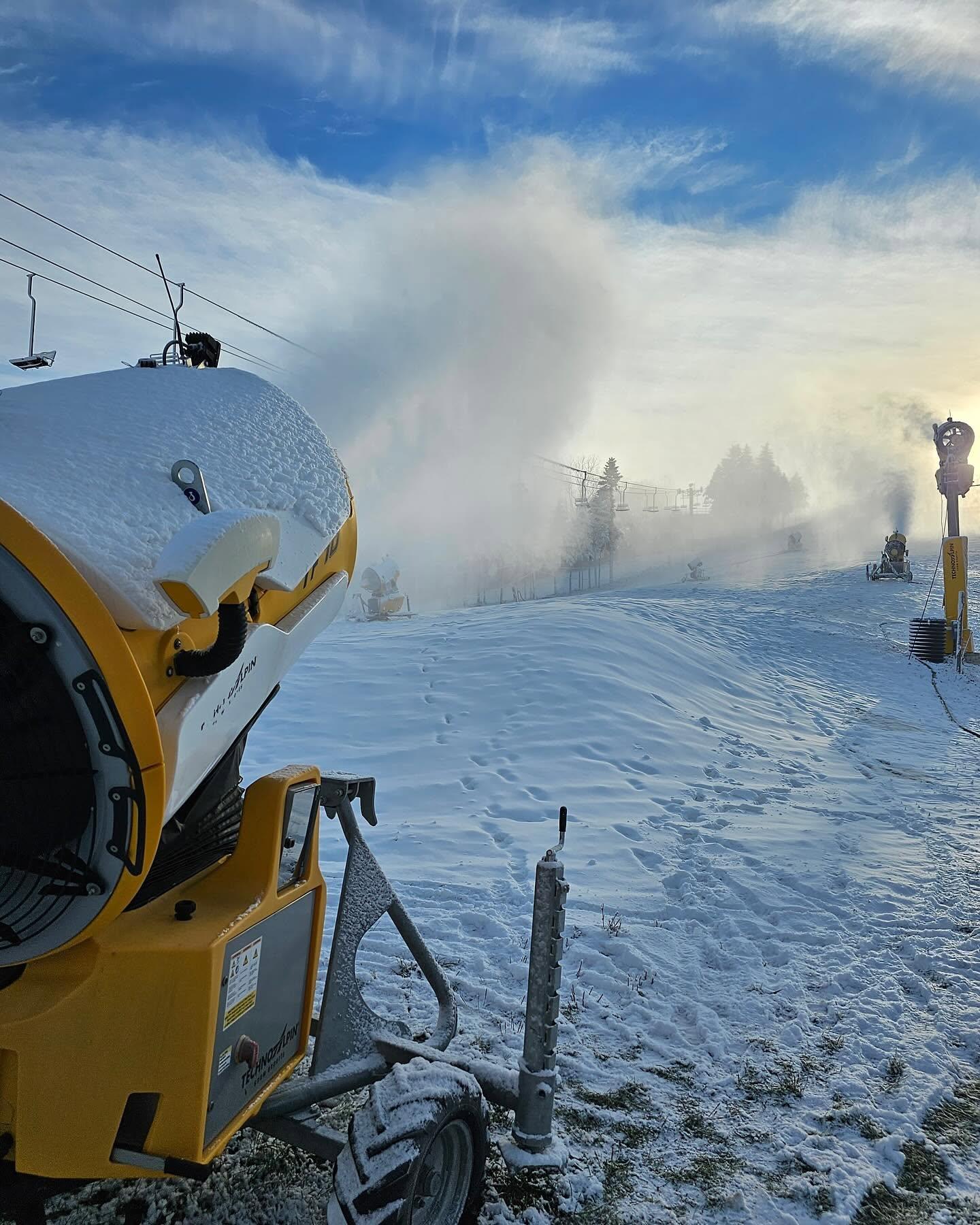 Snowmaking machine spraying mist across a ski slope beneath a blue sky