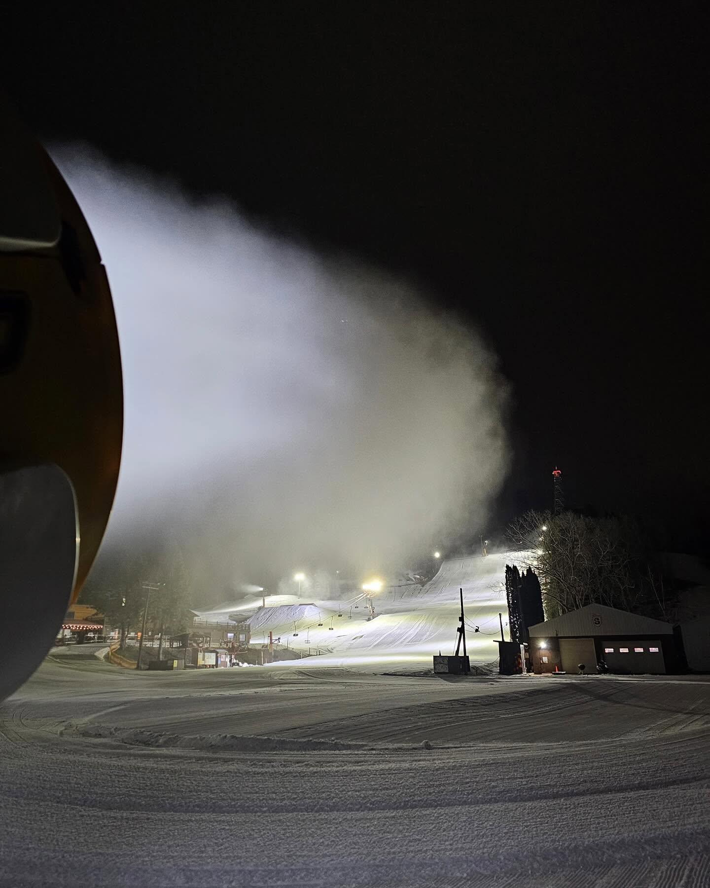 Snowmaking machine sprays a large plume of snow across a ski slope at night.