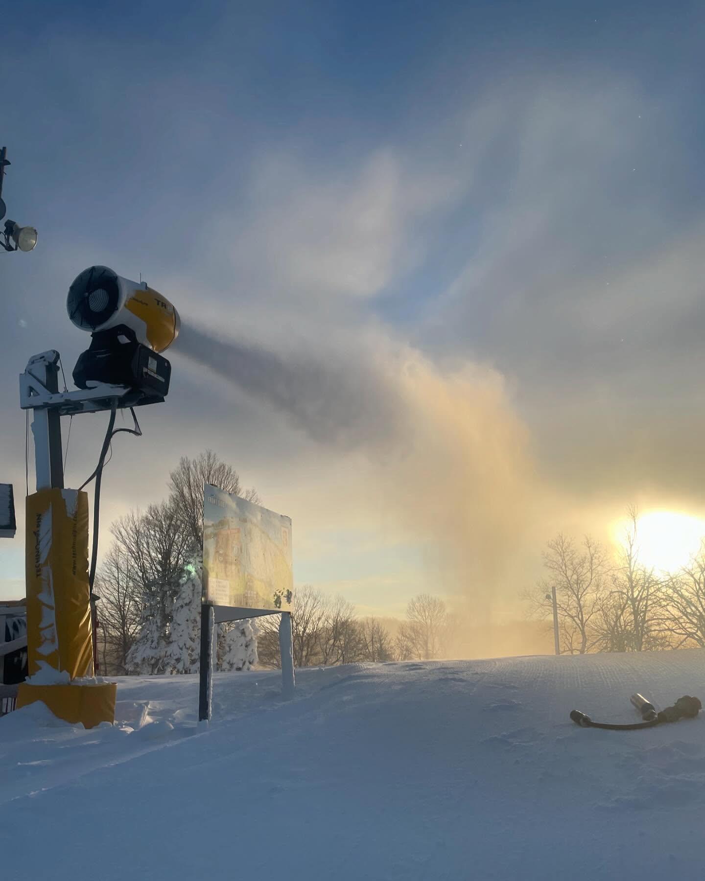 Snowy ski slope at sunrise with a snow cannon spraying mist.