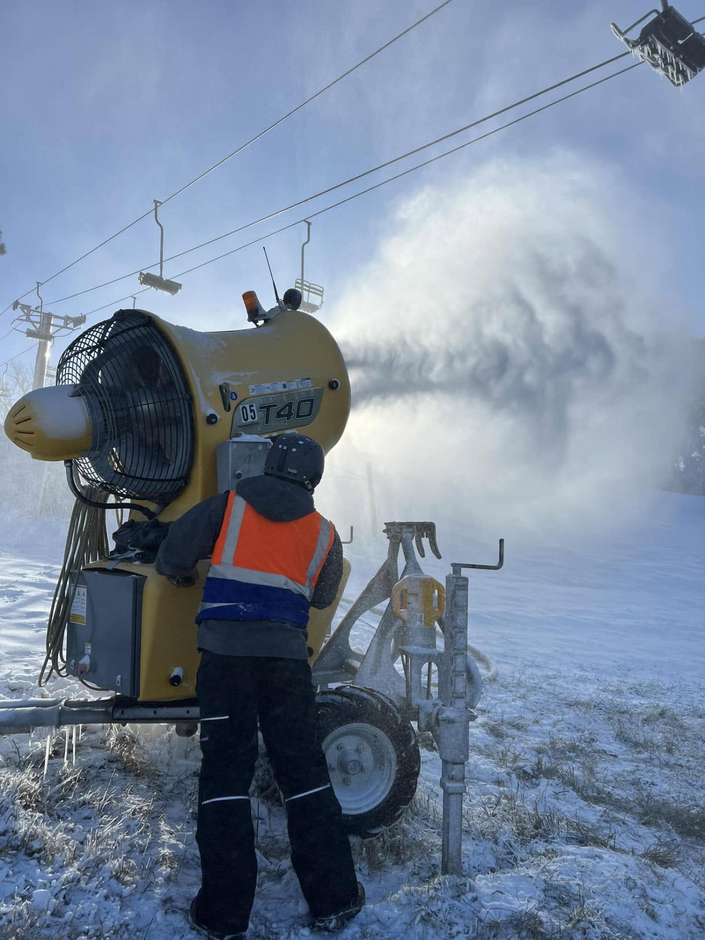 Worker operates a snowmaking machine on a frosty slope as mist billows behind.
