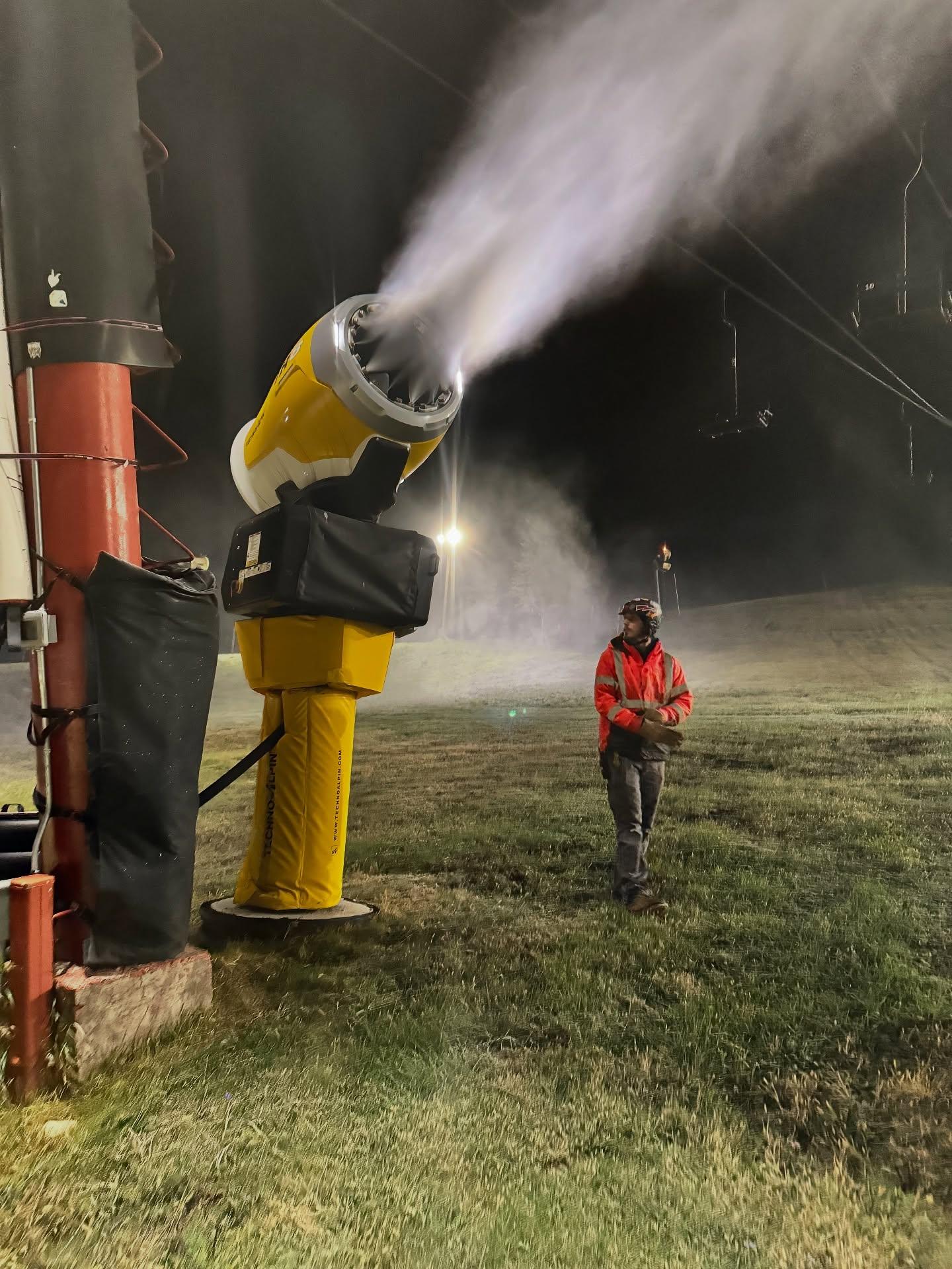 Worker in red jacket near a yellow snow cannon spraying mist at night