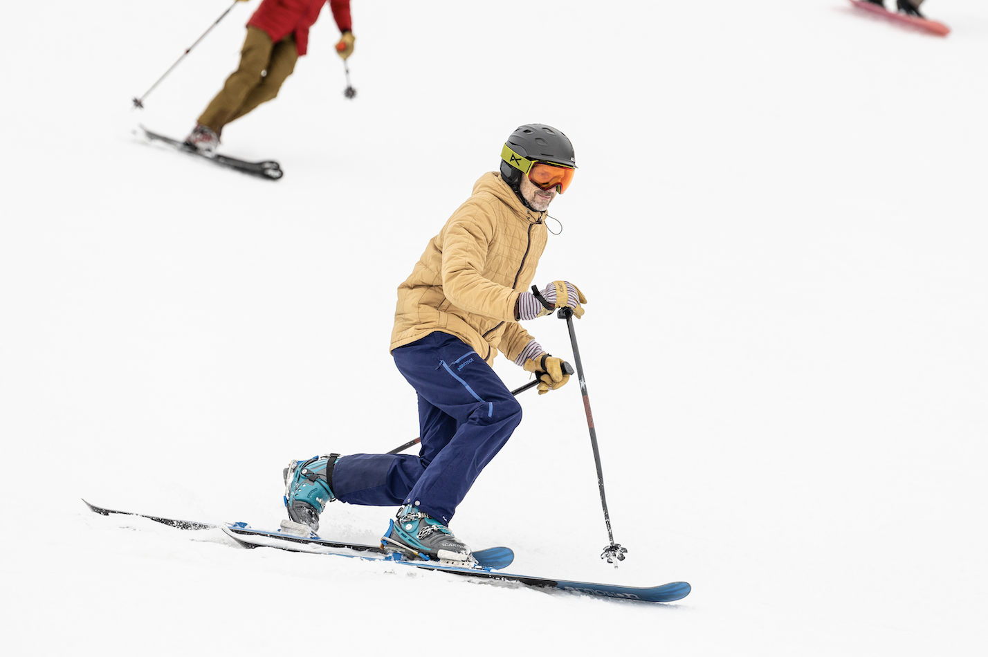 Skier in tan jacket and helmet carving on snowy slope.